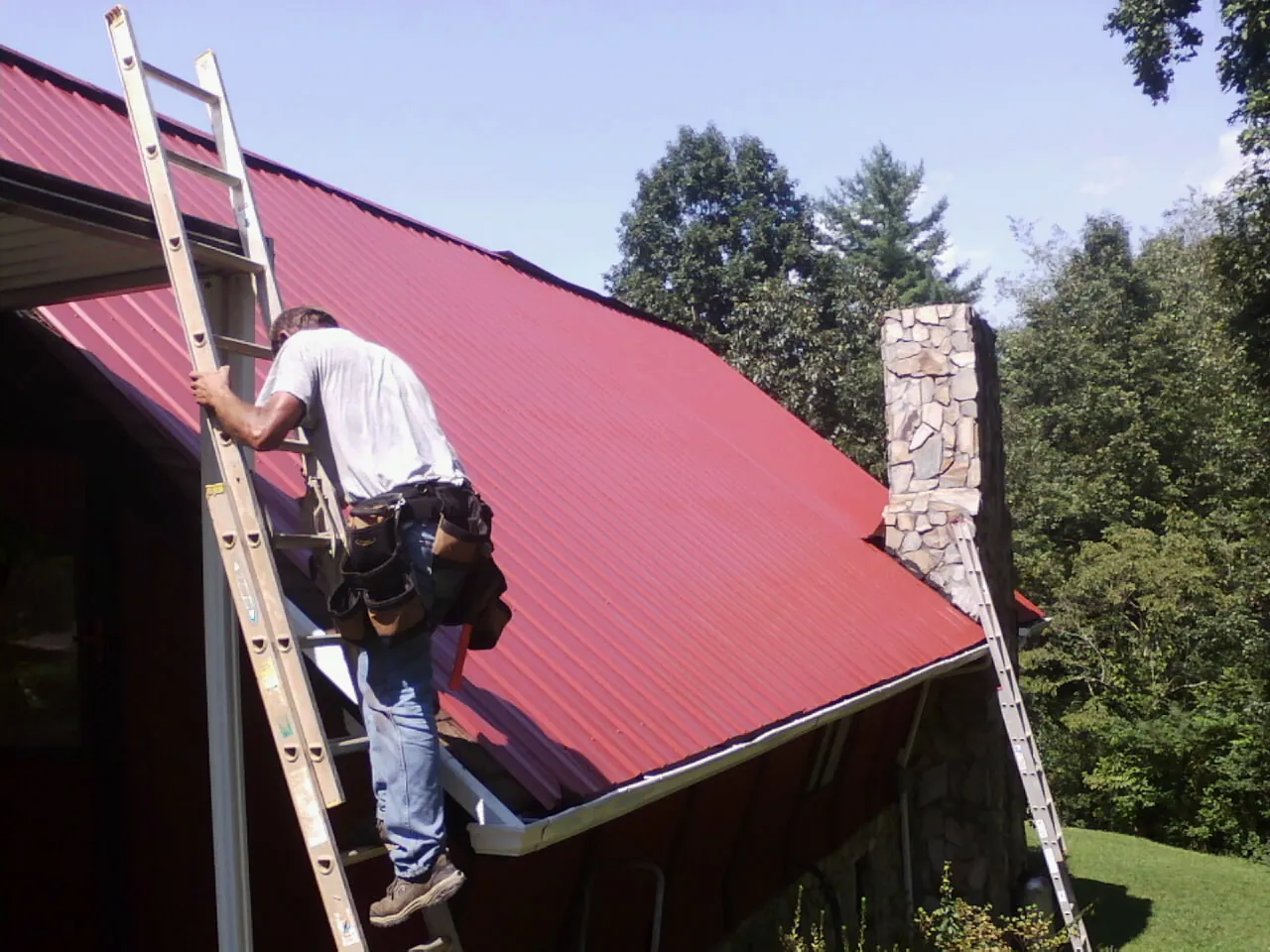Roofer on a ladder beside a red metal roof and stone chimney — Soffit & Fascia Repair by Roofing Brothers in Nellis Afb