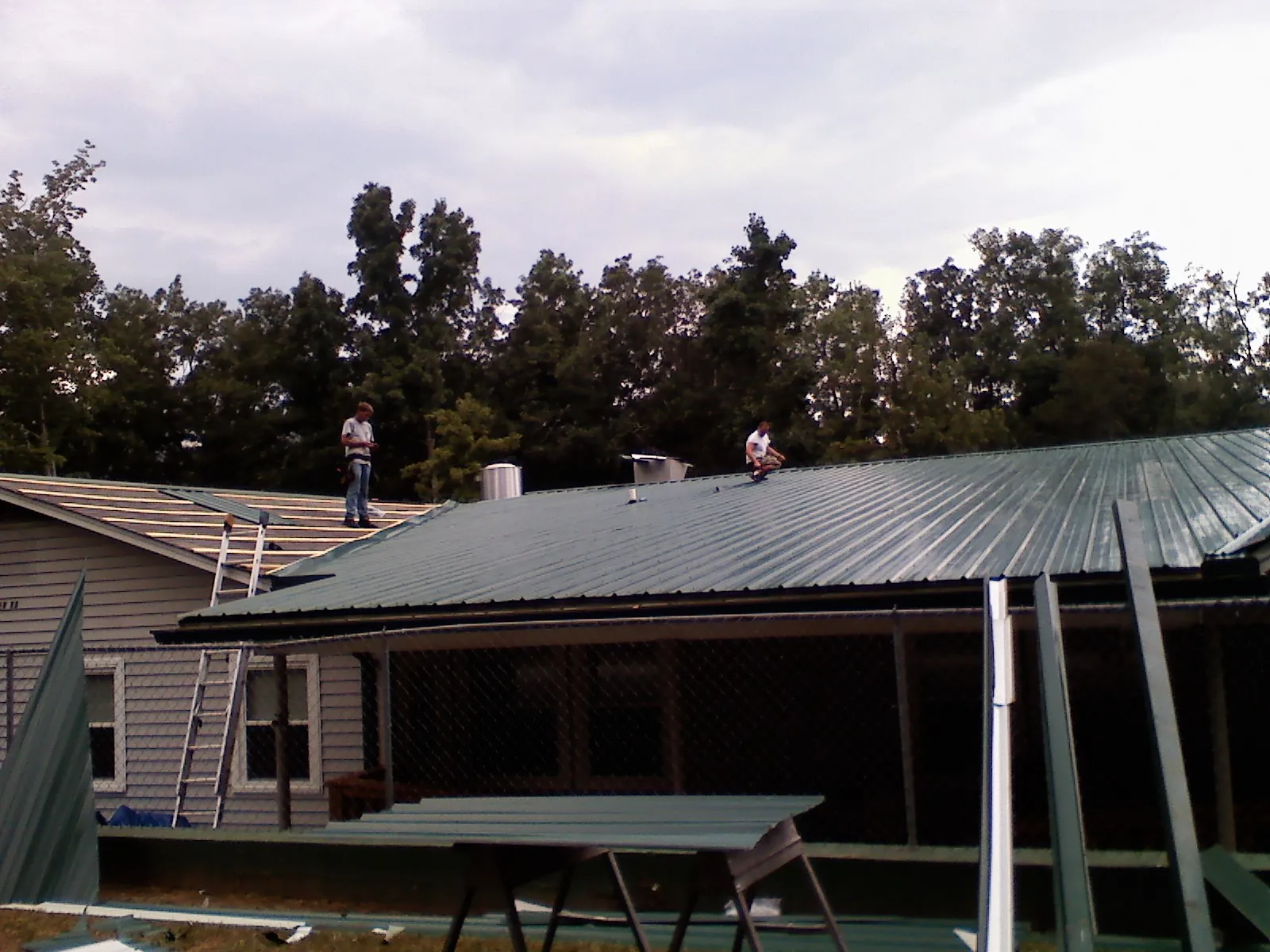 Two roofers installing green metal panels above a covered porch in Nellis Afb